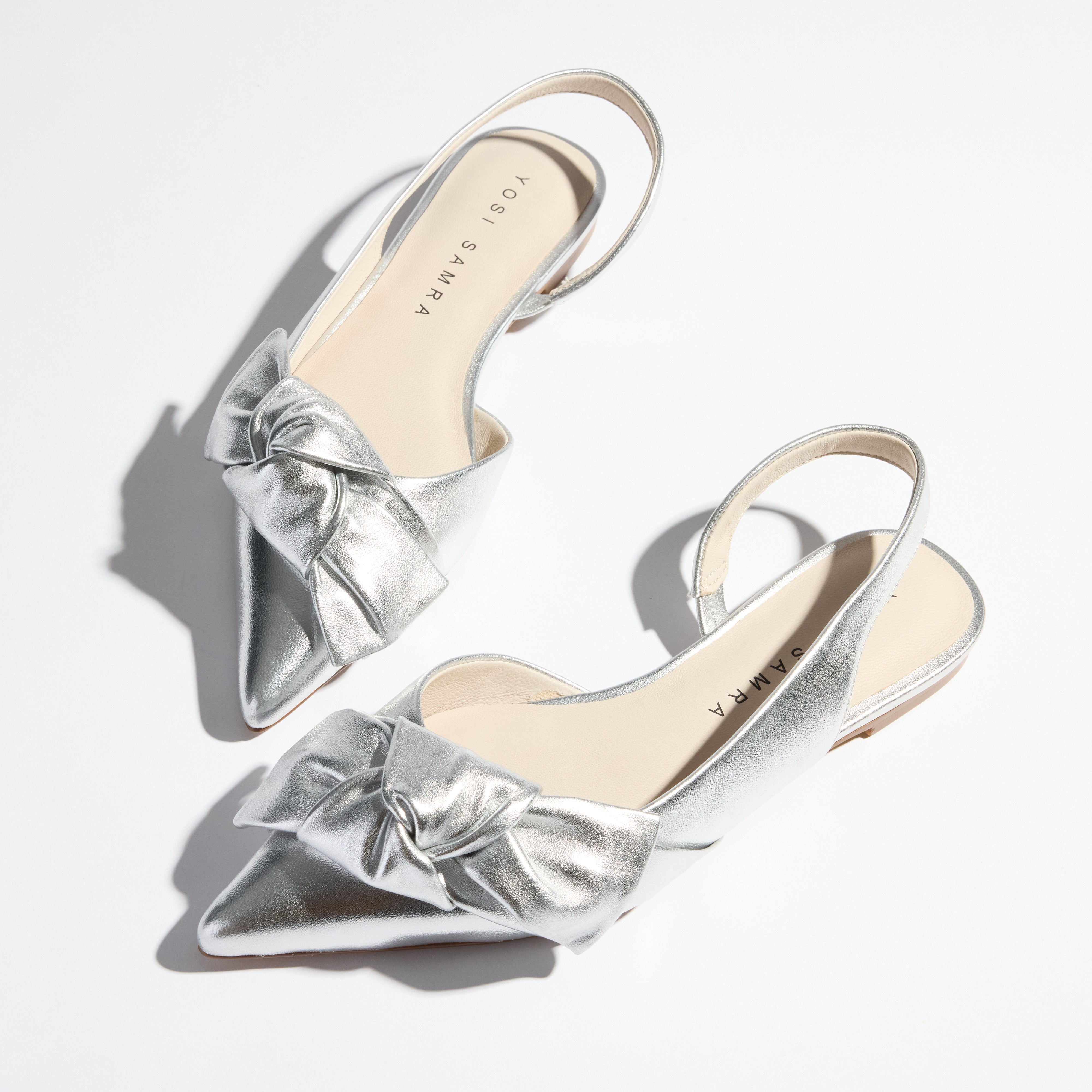 A pair of silver leather slingback flats with oversized bows on the pointed toes, displayed against a white background.